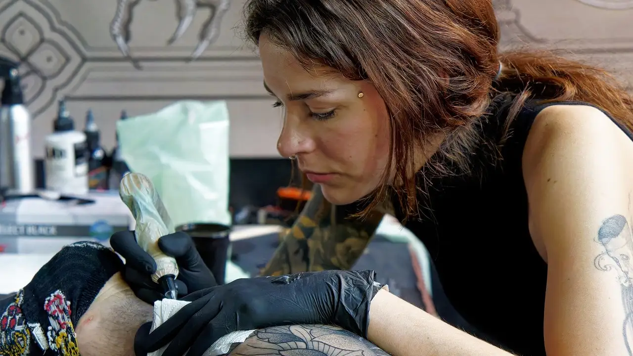 Tatuatoare la lucru pe un picior, mașina în mână, pielea întinsă cu mănușa